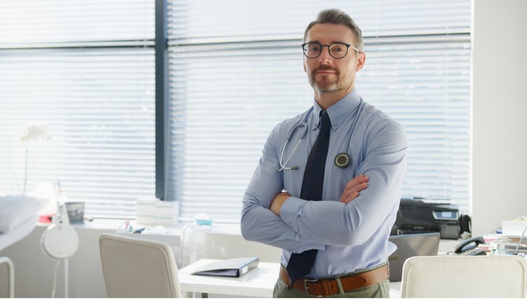 Mature Male Doctor Wearing Stethoscope Standing By Desk In Office